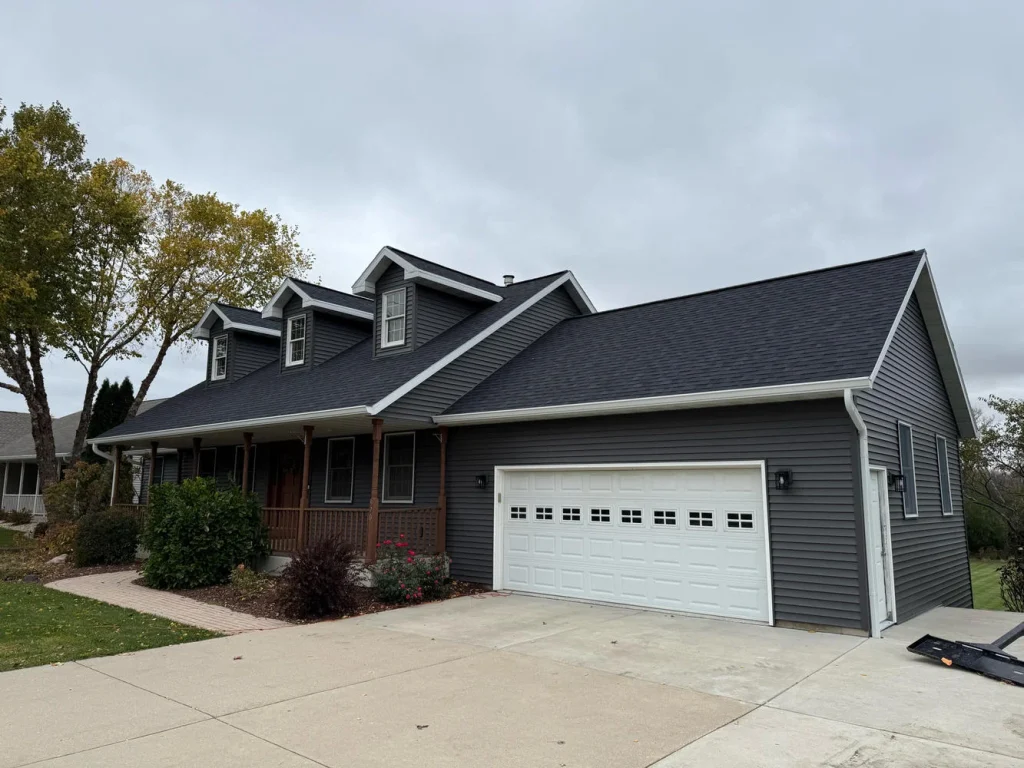 Gray house with dark gray shingles on a cloudy day, featuring white dormers, a porch, and a white garage door.